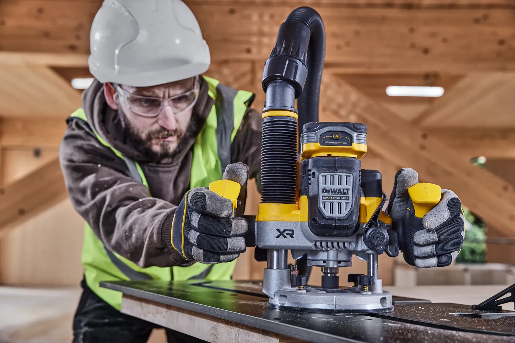 Worker uses a DEWALT XR router on wood, wearing safety gear in an indoor construction setting.