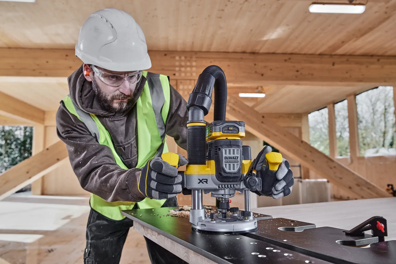 Worker uses DEWALT XR router DCW620NT on a metal surface in a wooden workshop, wearing safety gear.