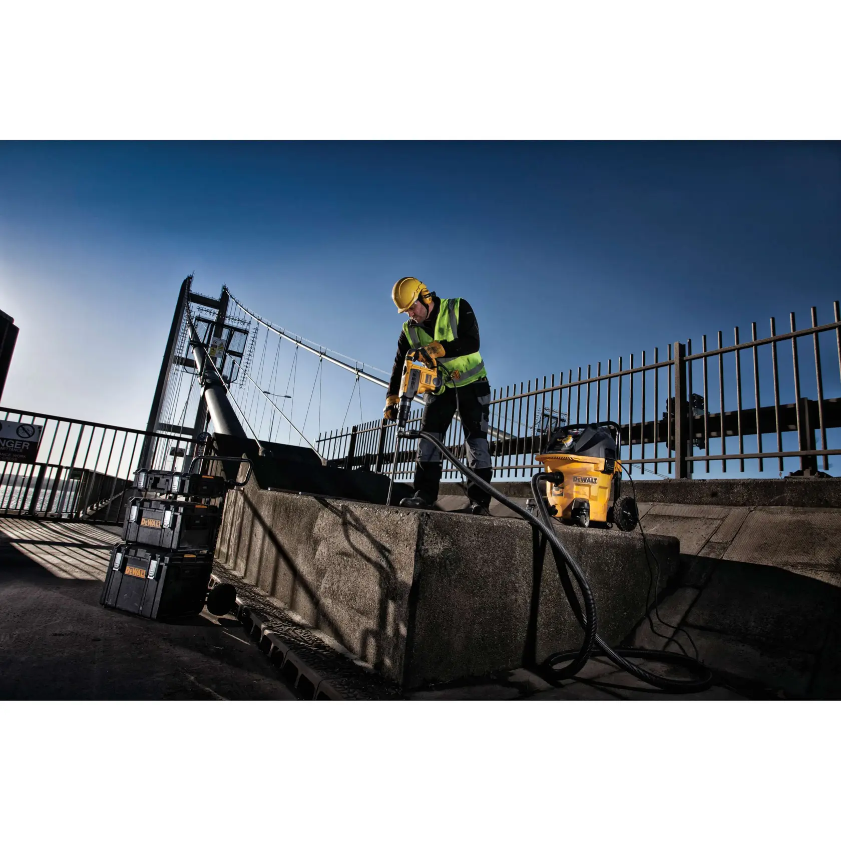 Trabajador usa herramienta y aspiradora DEWALT sobre hormigón, al aire libre, con chaleco y casco de seguridad.