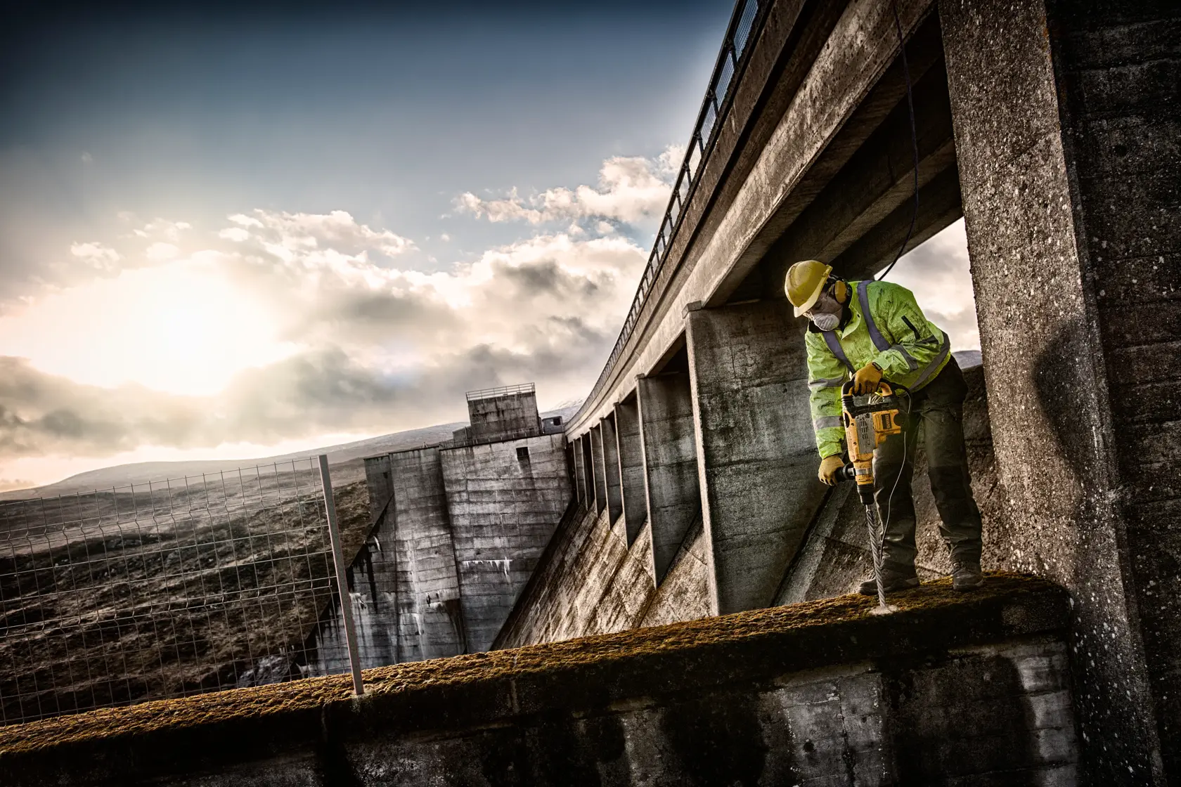 Arbeiter nutzt DEWALT Werkzeug zum Bohren von Beton am Staudamm, mit Schutzausrüstung auf einem Vorsprung.