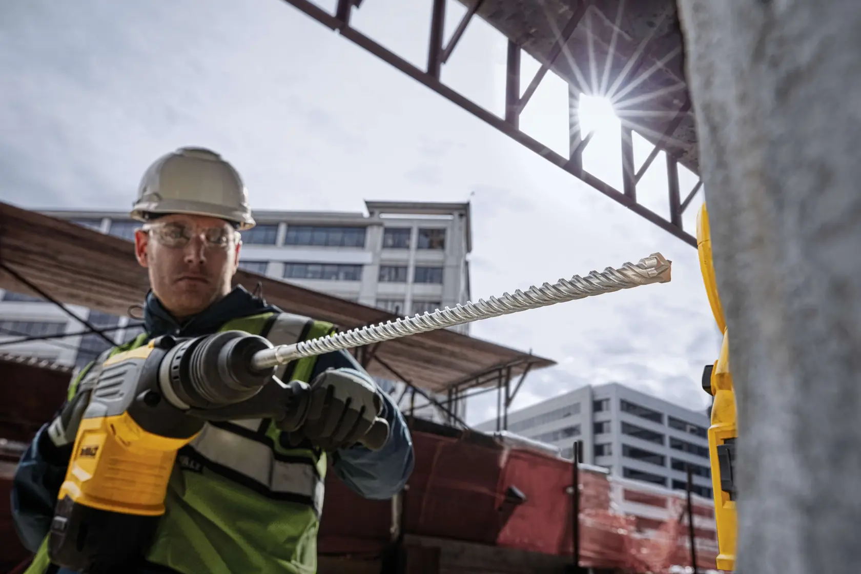 Arbeiter nutzt DEWALT-Bohrmaschine mit DT60825-Bit an Baustelle mit Gebäuden im Hintergrund.