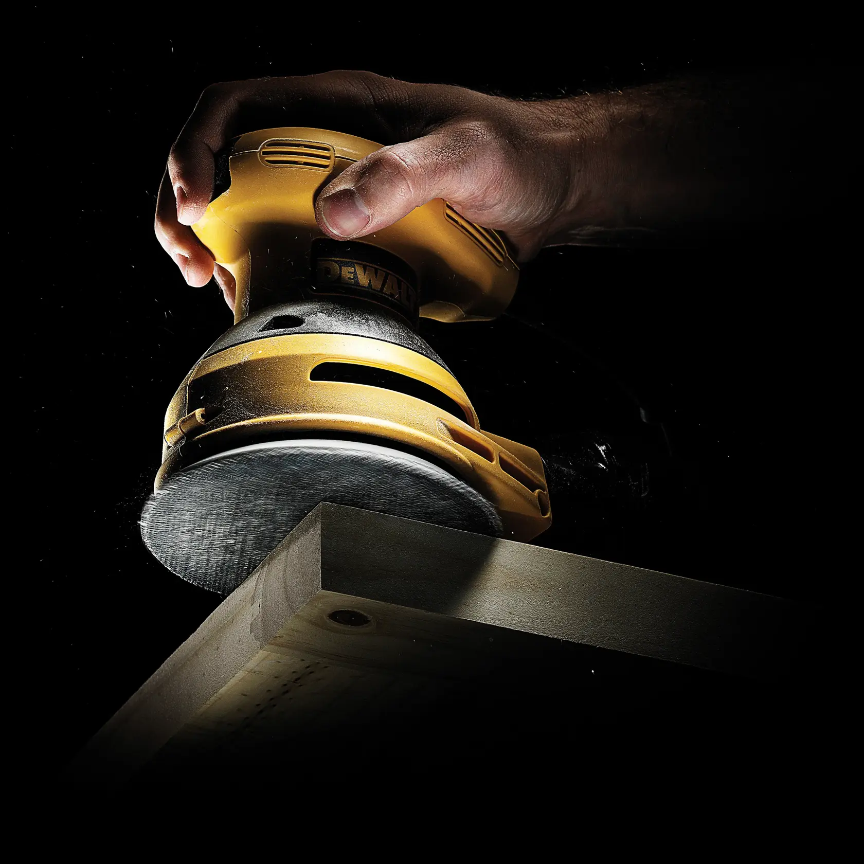 Hand using DEWALT sander on wooden surface against a black background.