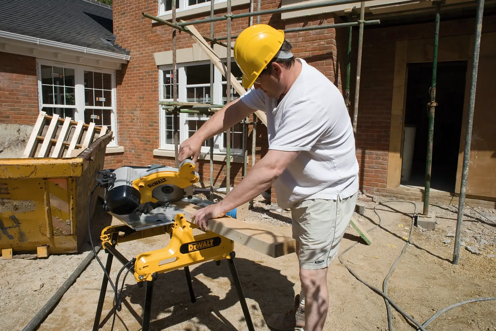 Person using a DEWALT DW743N saw to cut wood at a construction site with red brick buildings in the background.