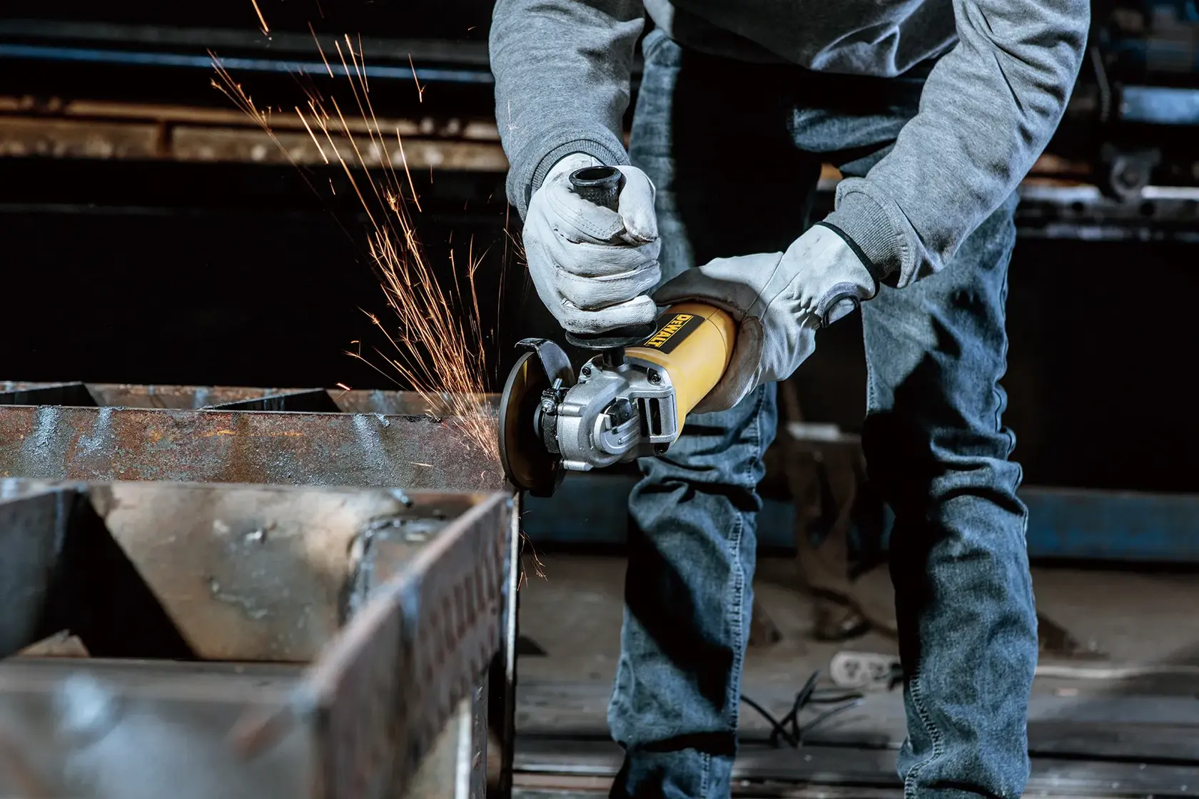 A person wearing gloves uses a yellow DEWALT angle grinder to grind a metal surface, producing sparks.