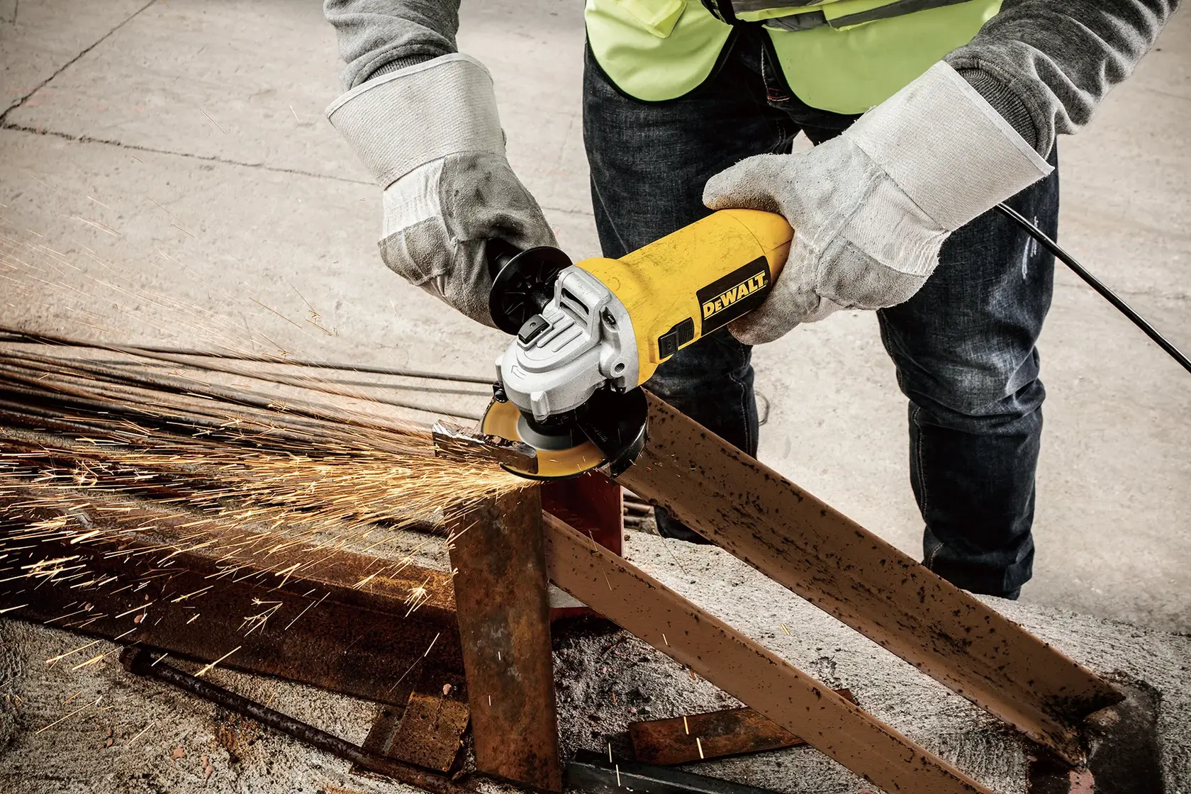 A person wearing safety gloves and a vest is using a yellow DEWALT angle grinder (model DWE4117) to cut through a metal beam, creating sparks.