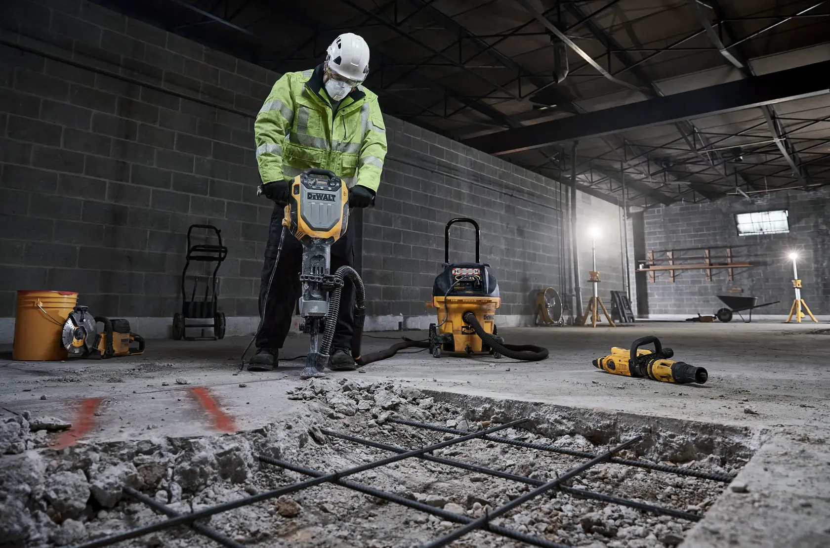 Worker uses a DEWALT jackhammer on concrete floor, surrounded by tools and construction equipment in an industrial setting.